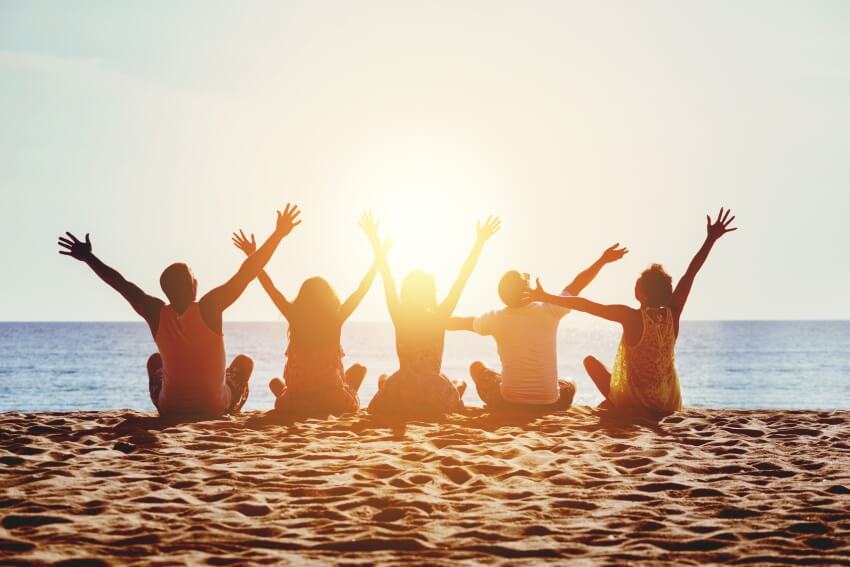 Fem jenter og gutter sitter på stranden med armene i luften og nyter solnedgangen