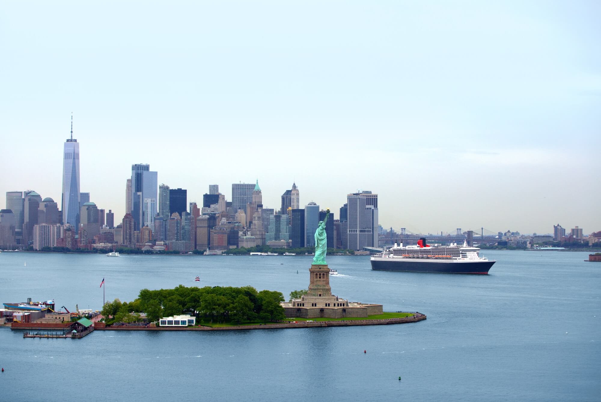 Cruiseskipet Queen Mary 2 seiler ut fra New York, med Frihetsgudinnen i forgrunnen og Manhattan i bakgrunnen. Foto.