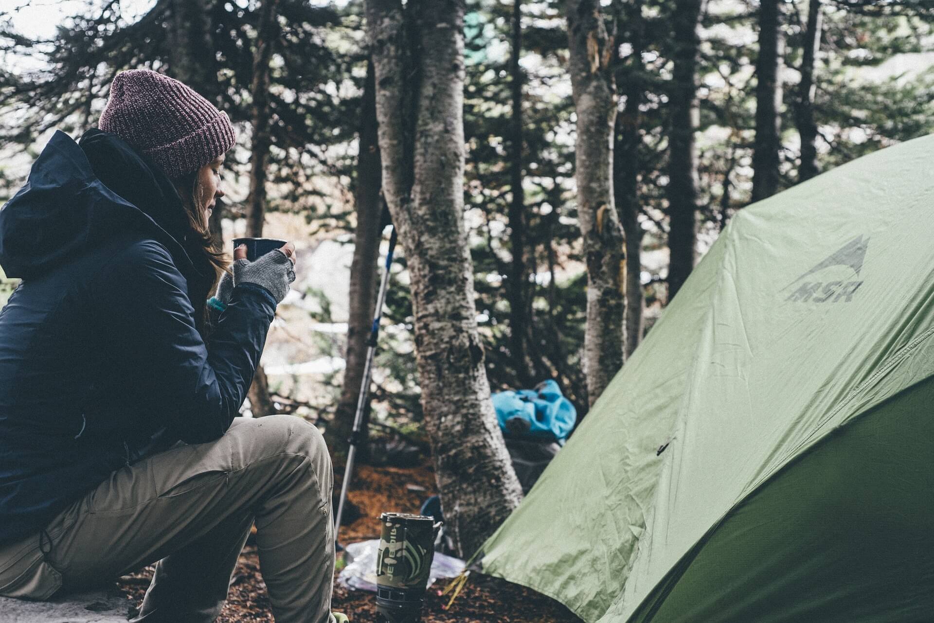 Jente sitter ute i skogen med varm drikke i hendene, foran et telt