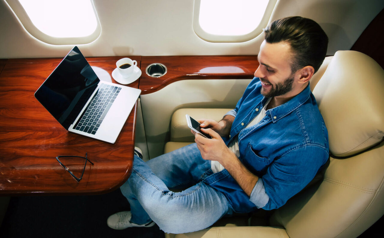 Mann ombord et privat jetfly med pc foran seg mens han taster på mobiltelefonen
