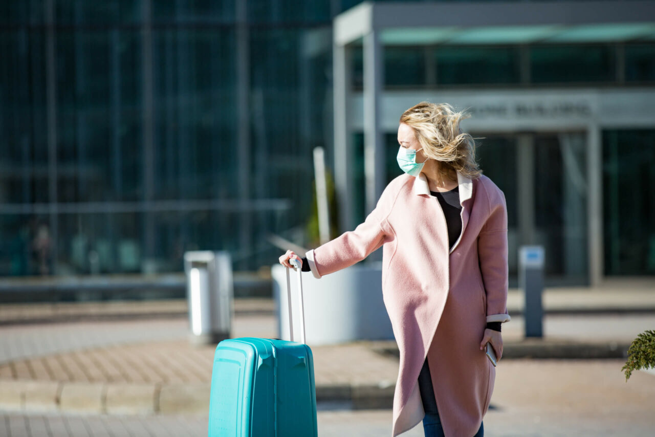 Kvinne som venter utenfor en flyplass med koffert og munnbind