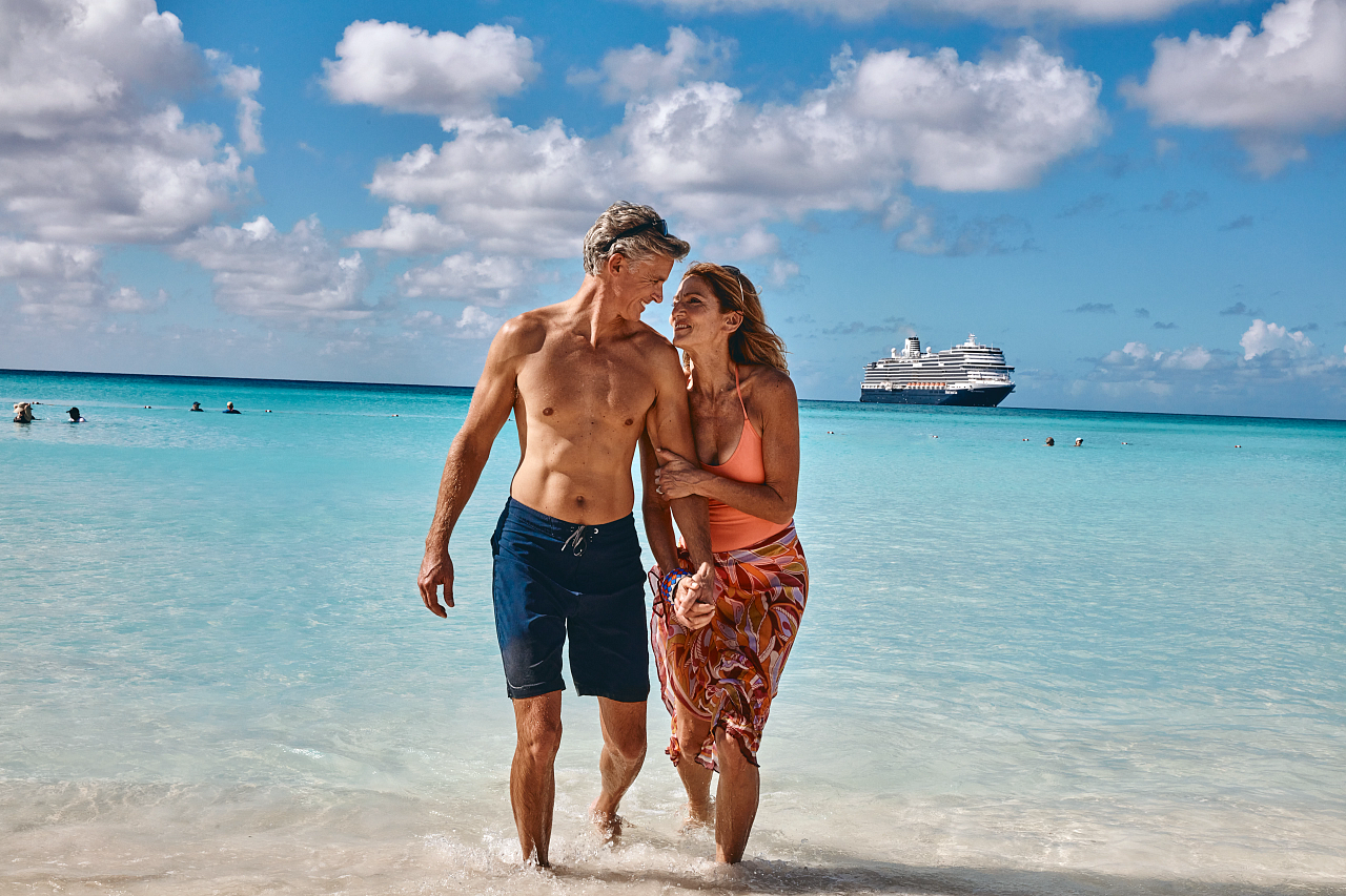 Par går hånd-i-hånd i vannkanten på en strand i Karibia, med et cruiseskip i bakgrunnen. Foto.