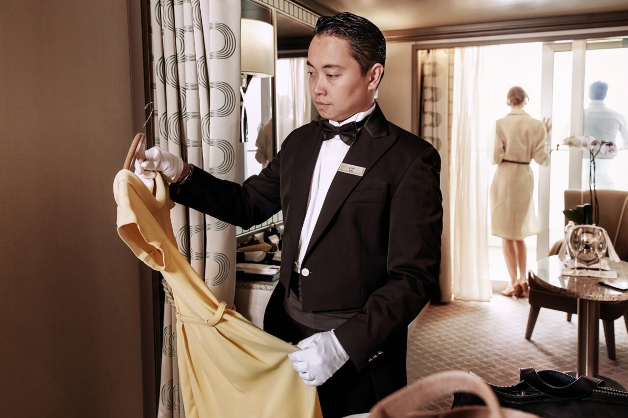 Butler henger opp klær i en suite om bord på Silversea, med to gjester i bakgrunnen på balkongen. Foto.