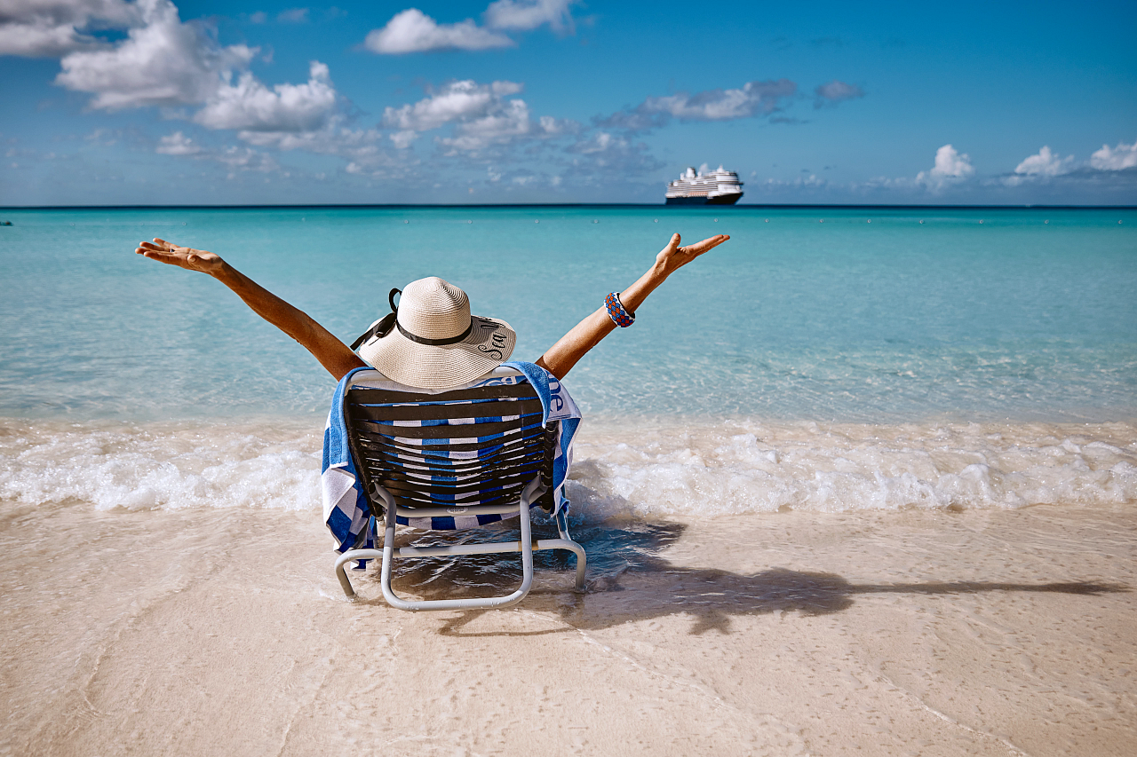 Dame med solhatt sitter i en solseng på stranda, med et Holland America Line-skip i bakgrunnen.