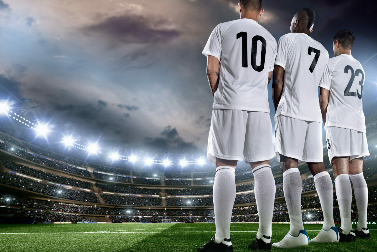 Tre fotballspillere står på fotballbanen med ryggen mot kamera