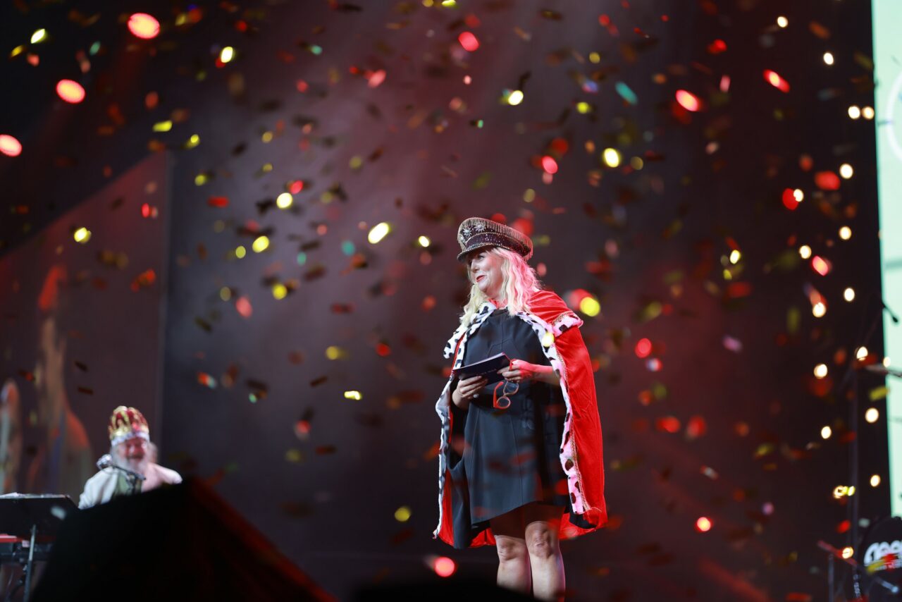 Kvinne står på scenen kledd i kappe og hatt, mens det regner konfetti. Foto