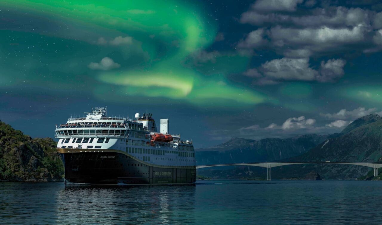 Cruiseskip ligger til sjøs med dansende grønt nordlys over seg. Foto