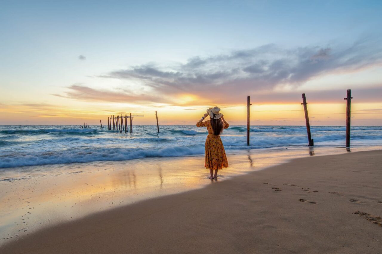 En dame med ryggen til på en sandstrand i solnedgangen