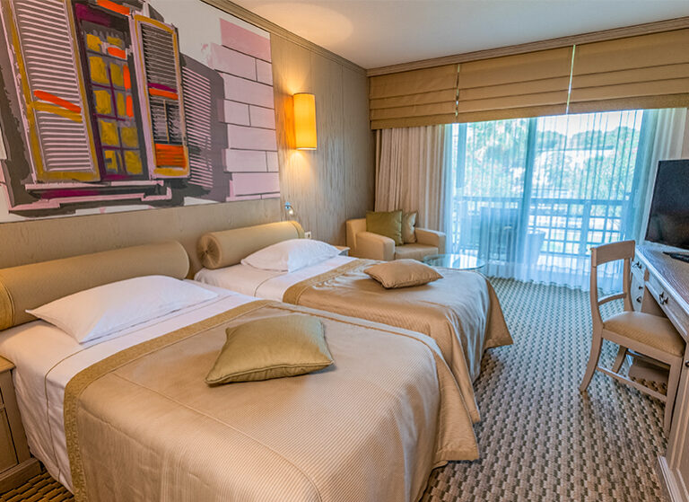 To senger i beige på standard rom, med balkong og skrivebord, stol og tv. Foto