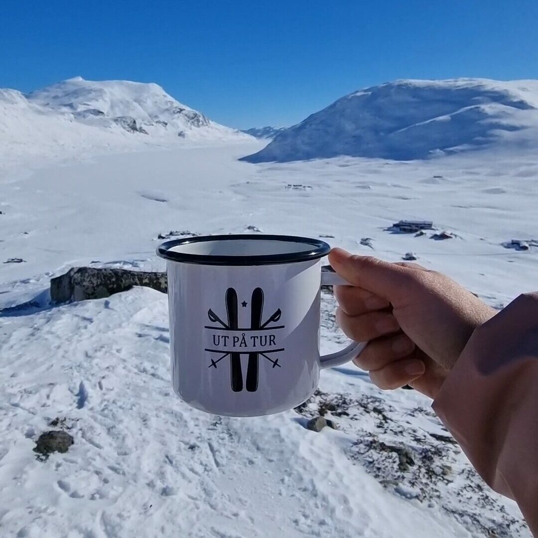 Hvit kopp med teksten "ut på tur" i vinterlandskap. Foto