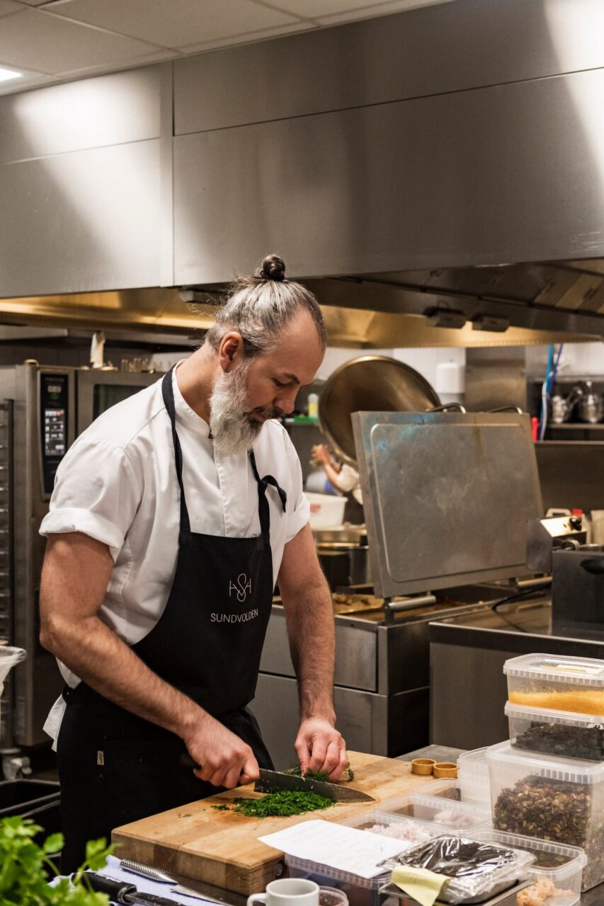 Mann i kokkeuniform står på et kjøkken og hakker grønsaker. Foto
