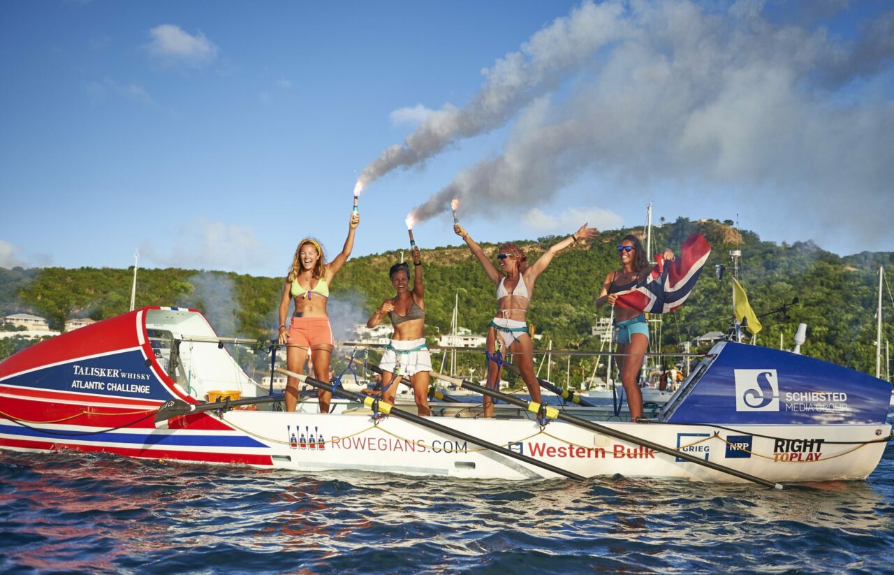 Fire kvinner om bord en robåt ser inn i kamera mens de står med armene i været. Foto