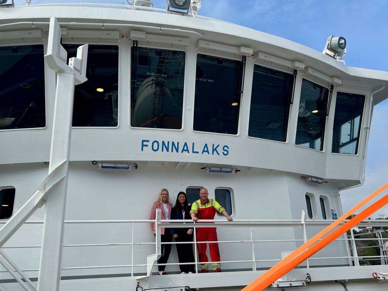 Tre mennesker står om bord et skip og smiler inn i kamera. Foto