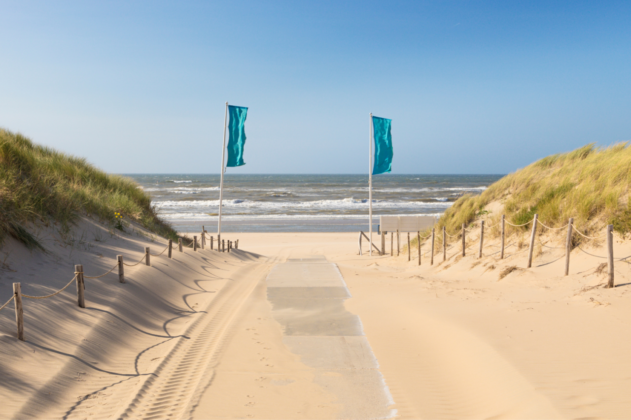 En flott sandstrand mot havet. Det vaier to blå flagg som ønsker folk velkommen til stranden. Foto