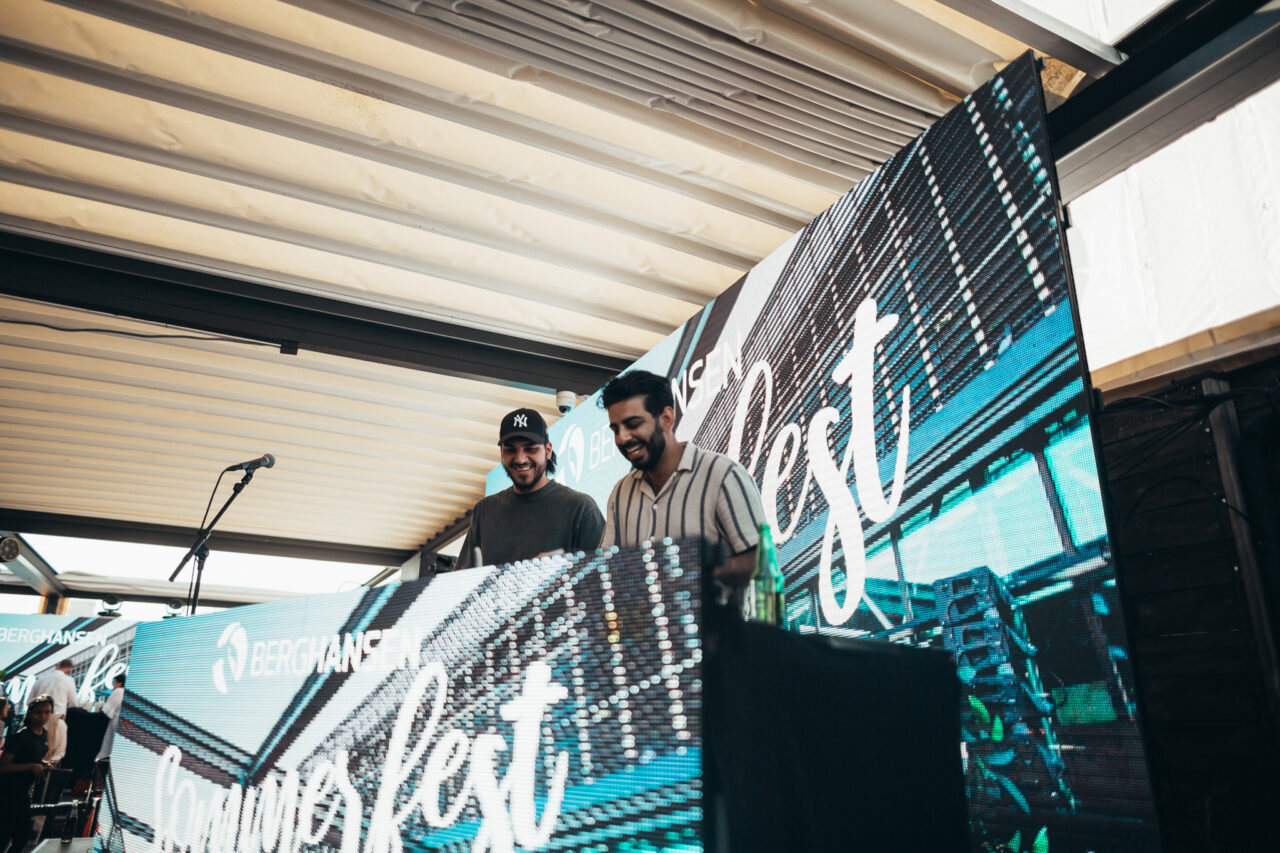 To menn står bak en DJ-pult og smiler. Foto