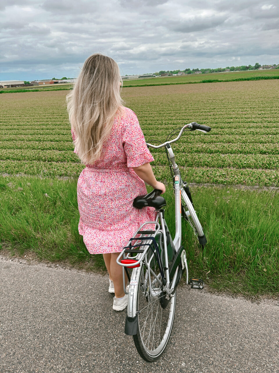 Jente i rosa kjole står med en sykkel foran en grønn åker. Foto