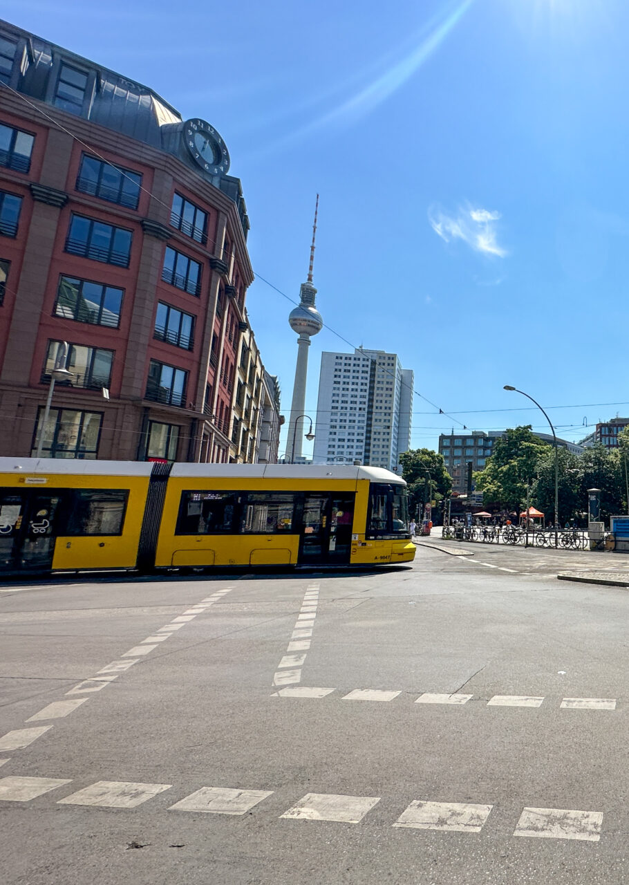En gul trikk som kjører over et veikryss i Berlin med TV-tårn i bakgrunnen. Foto