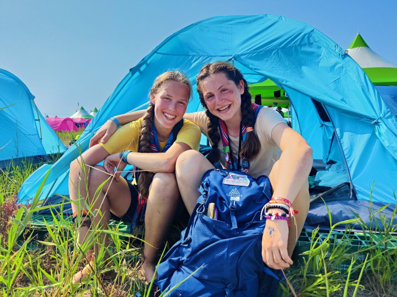 To speidere sitter foran en oppslått telt og smiler inn i kamera. Foto