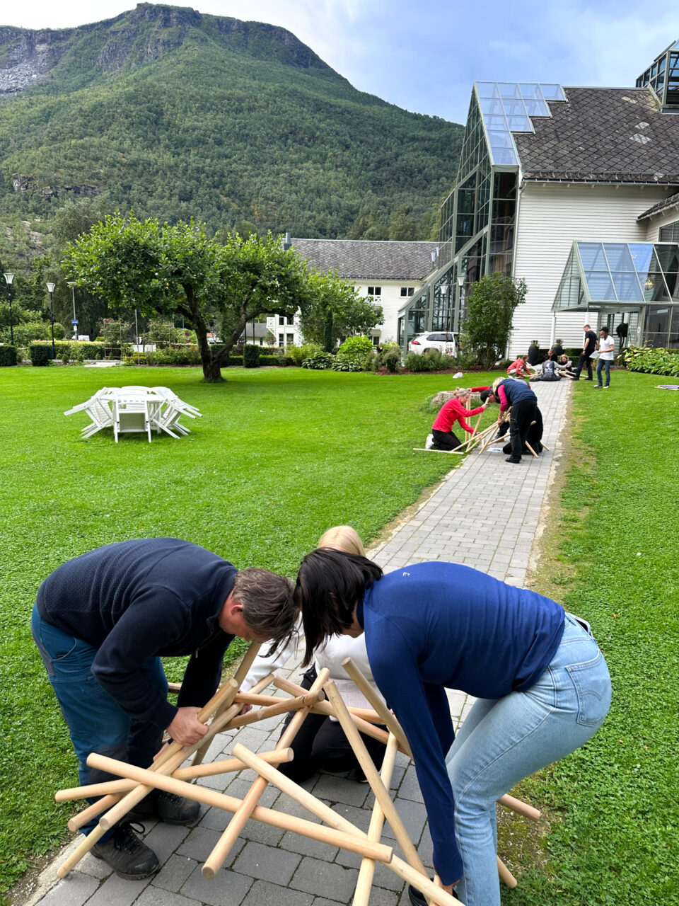 Tre personer som bygger noe med trepinner utendørs i en vakker hage. Foto