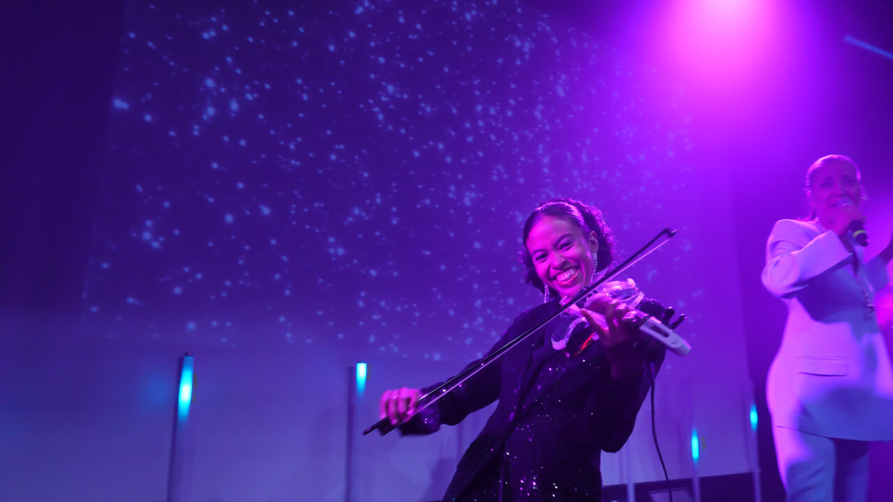 Kvinne spiller fiolin mens hun ser inn i kamera og smiler. Foto