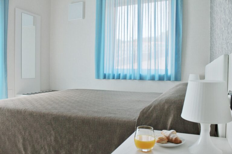 Dobbelseng i brunt sengeteppe, hvite vegger og et vindu med lyseblå gardin i bakgrunnen. Foto