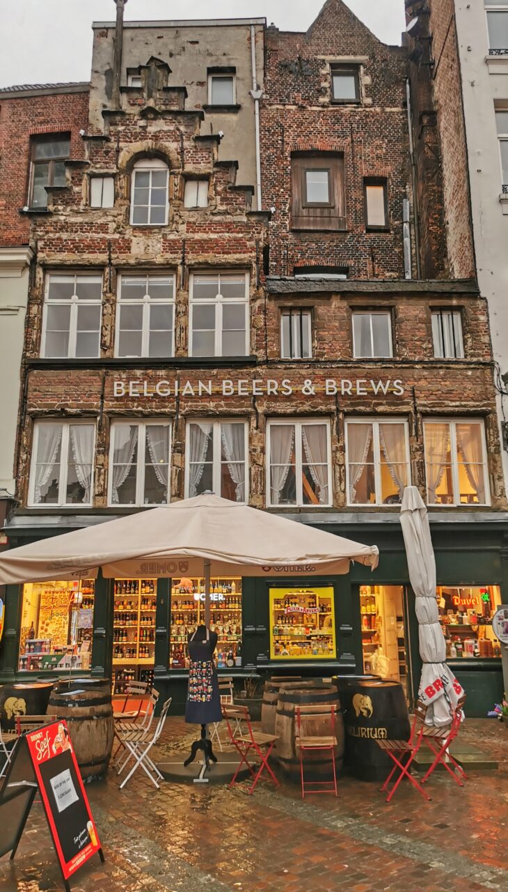 Et høyt bygg i murstein med sitteplasser og parasoll utenfor. På veggen står det "Belgian Beers & Brews". Foto