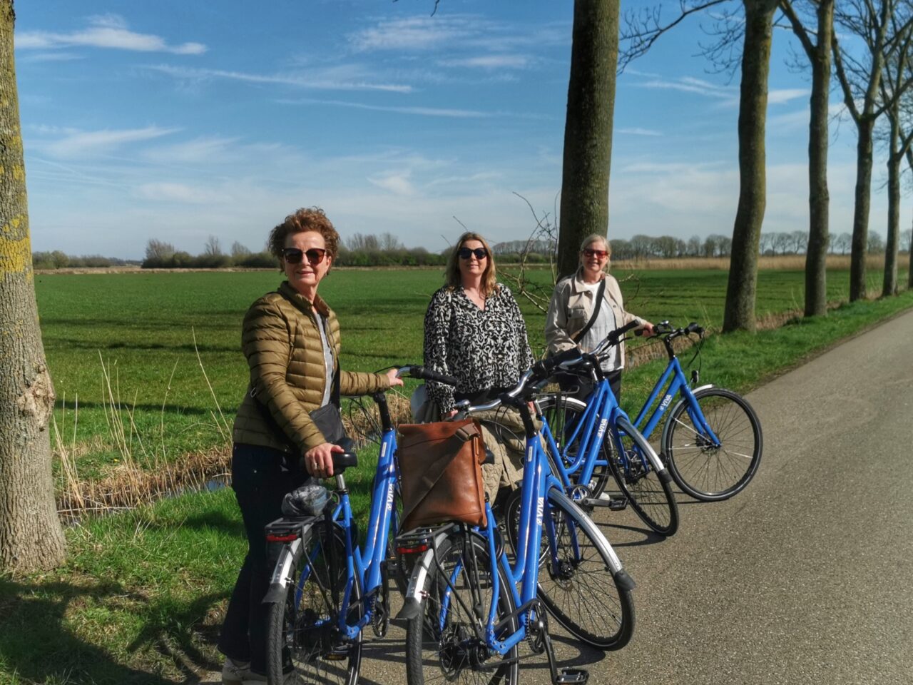 Tre damer står foran en grønn slette og landevei med hver sin blå sykkel og smiler mot kamera i Nijmegen, Nederland. Foto
