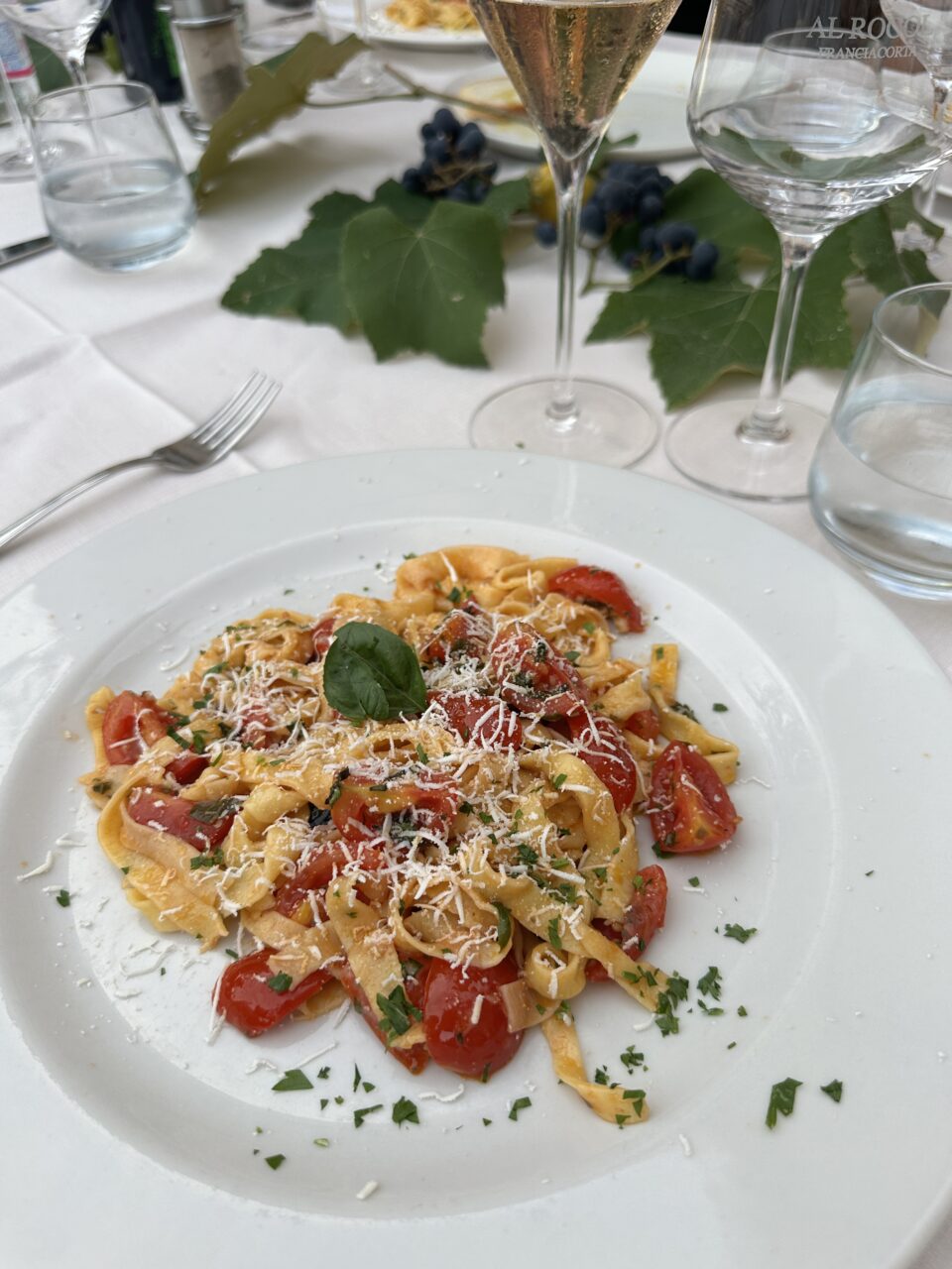 Nærbilde av en hvit tallerken med en pastarett bestående av pasta, tomater og basilikum på et dekket bord. Foto