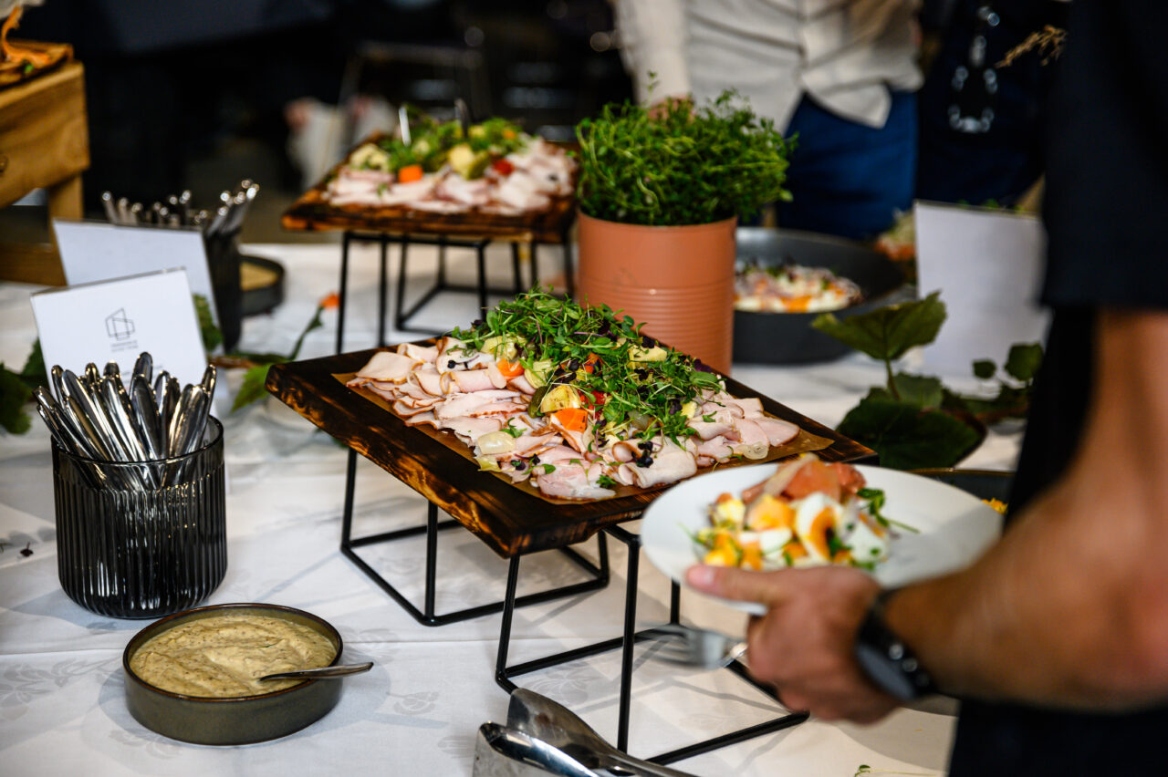 Buffetbord med et utvalg av matretter, inkludert serveringsfat med oppskåret kjøtt pyntet med friske urter og grønnsaker. En person holder en tallerken med mat i høyre hjørne av bildet. Foto