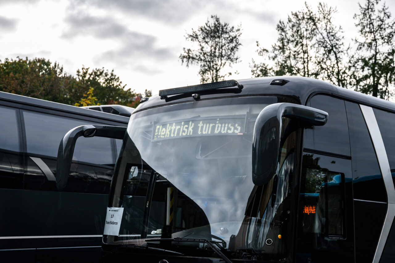 Fronten av en elektrisk turbuss med teksten 'Elektrisk turbuss' synlig på en digital skjerm i frontruten. Bussen står parkert utendørs, omgitt av andre busser og trær i bakgrunnen under en overskyet himmel. Foto