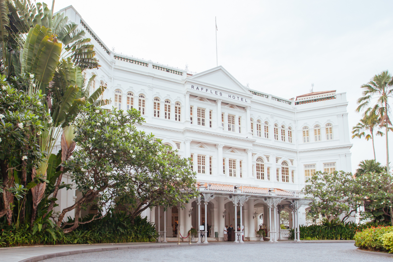 Fasaden til det ikoniske Raffles Hotel i Singapore, omgitt av frodige palmer og grøntområder. Foto